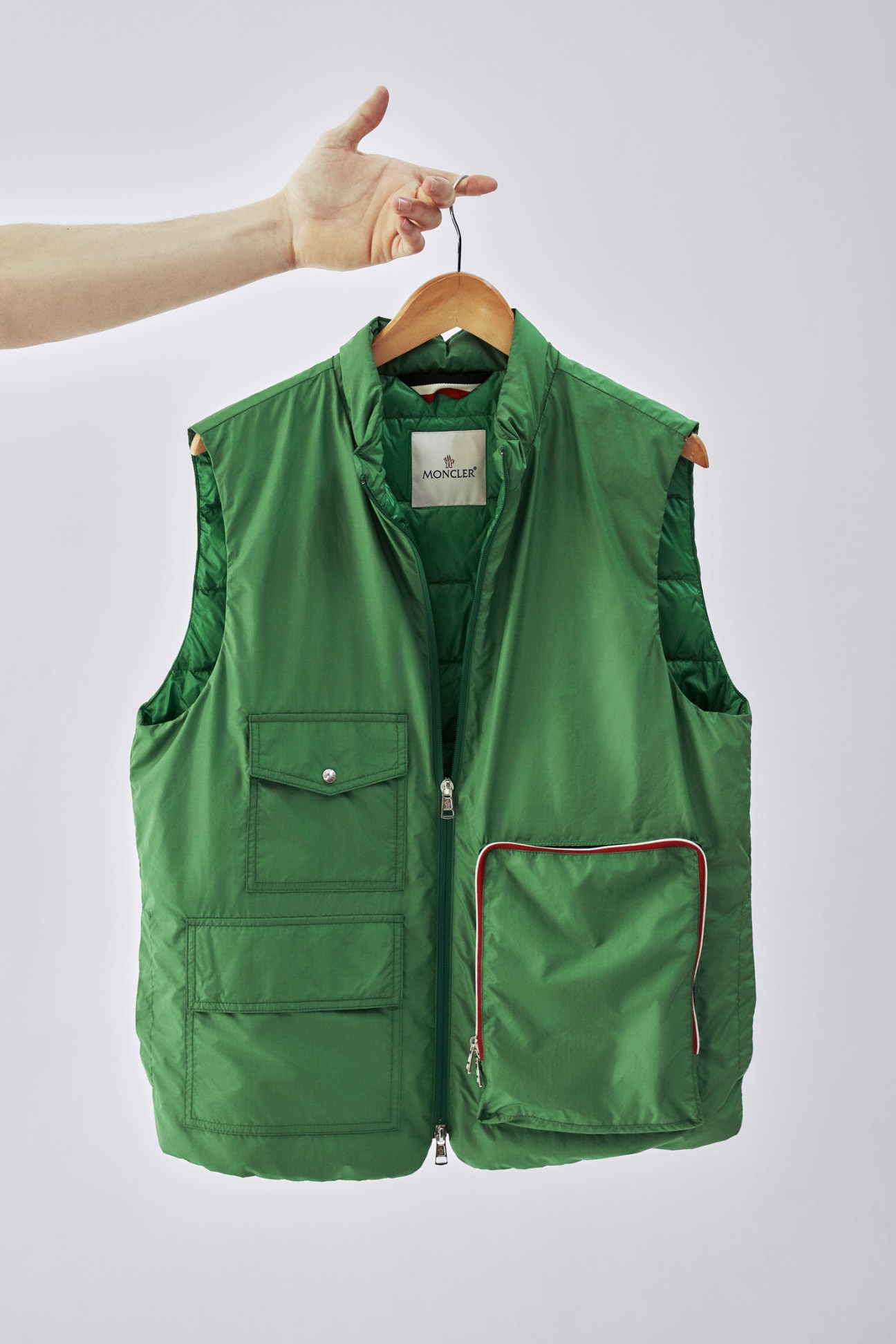 Hand holding wooden hanger with green Moncler down vest against white background.