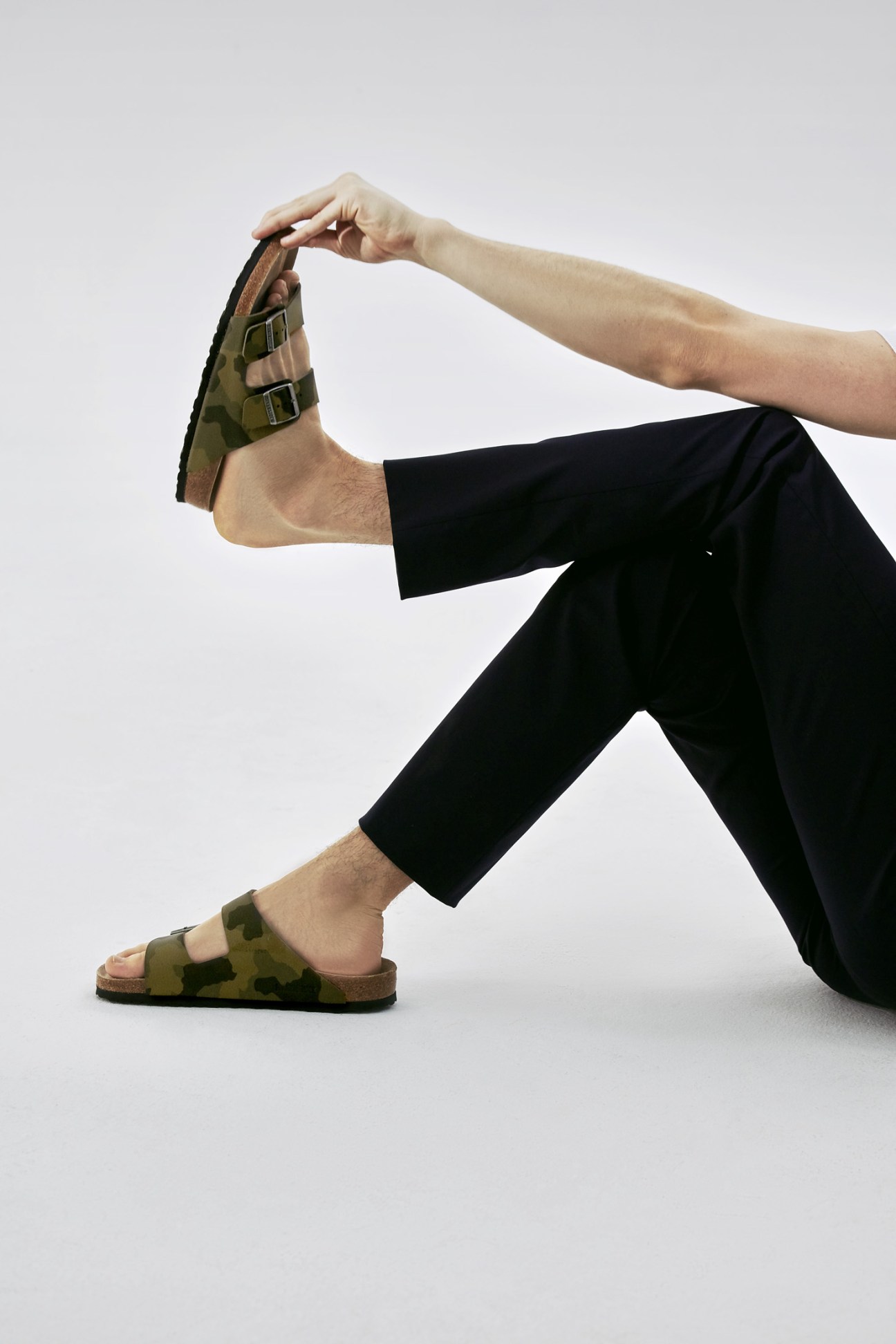 Person wearing black trousers and camouflage Birkenstock sandals, holding up a brown shoe with buckle details.