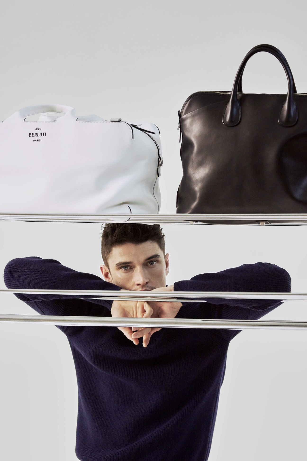 Model wearing navy jumper displayed behind shelves with white Berluti bag and black Valextra bag.