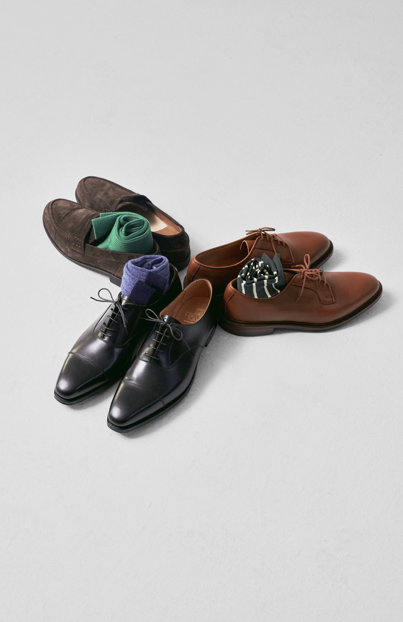 Three pairs of shoes with colorful socks: dark suede loafers, black oxfords, and brown derbies arranged on white background.