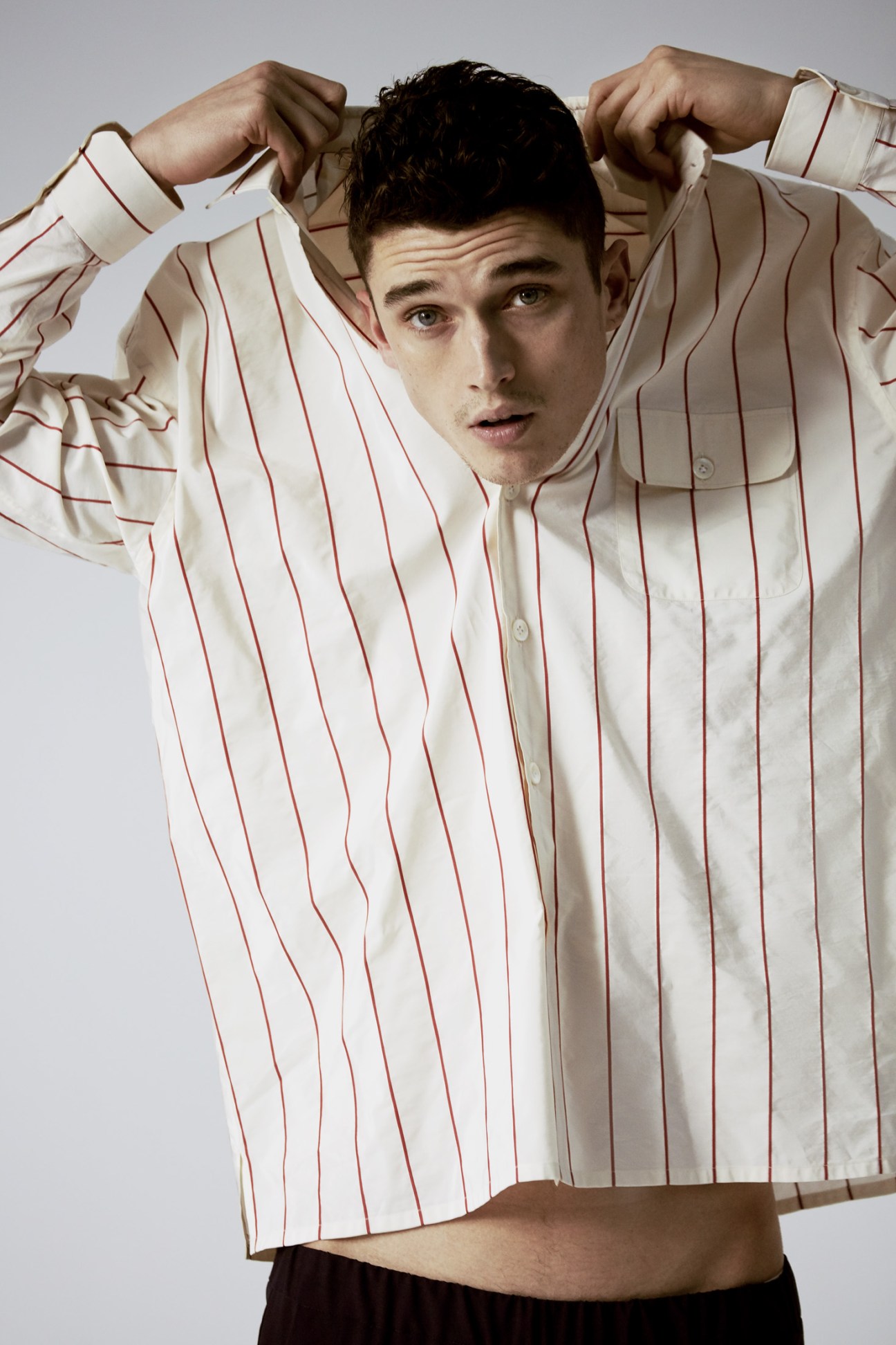 Man wearing cream shirt with red pinstripes by Pal Zileri, adjusting his dark hair against a grey background.