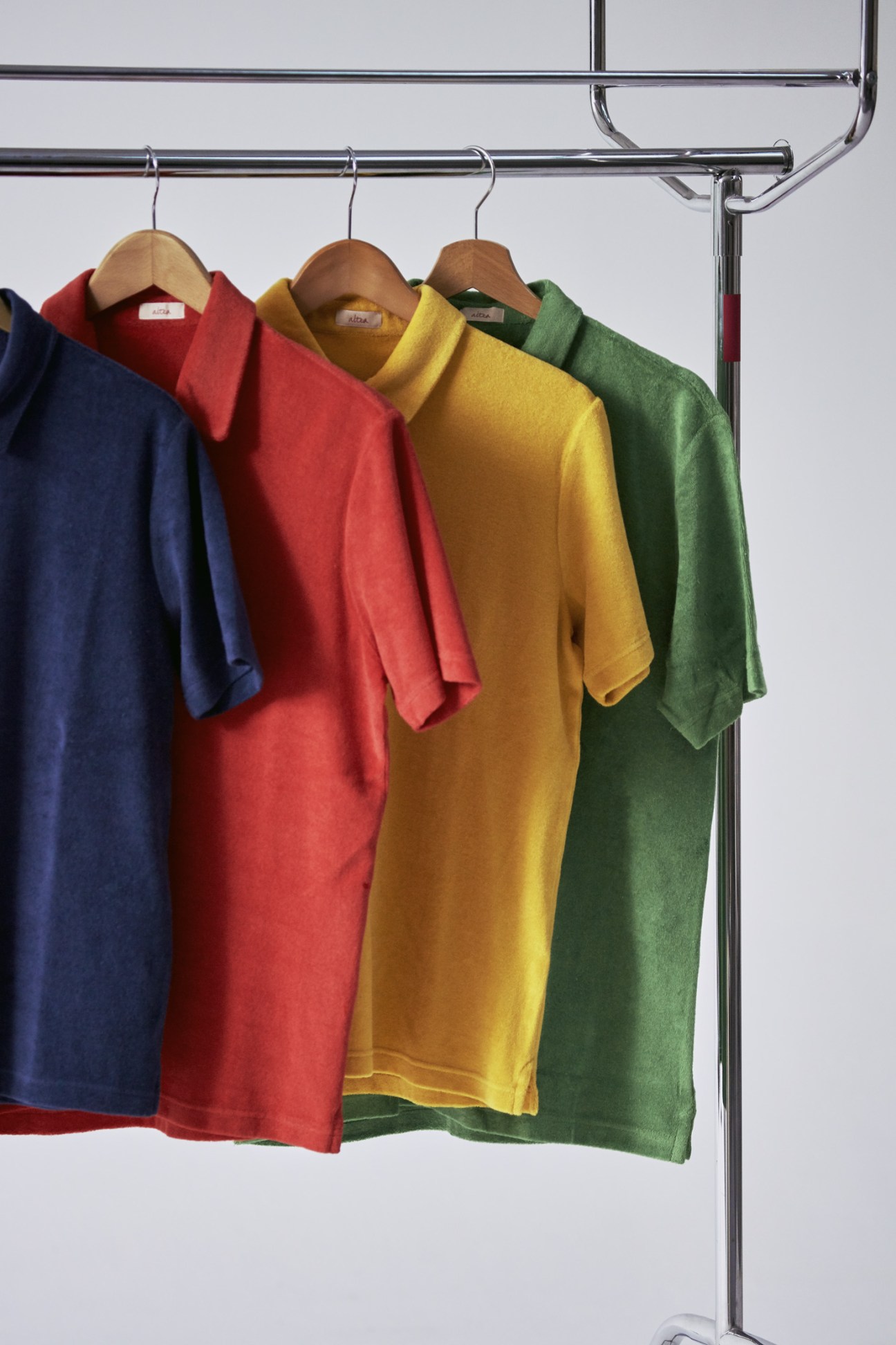 Polo shirts by Altea in navy, red, mustard yellow, and green hanging on wooden hangers from a chrome rail.