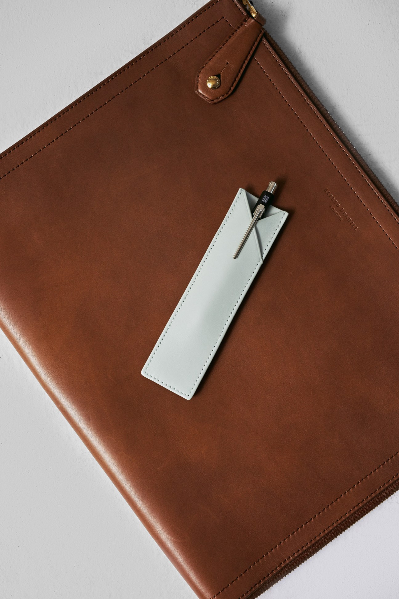Brown leather portfolio case by Dunhill with white pen case by Treuleben and pen by Caran d'Ache on white background.