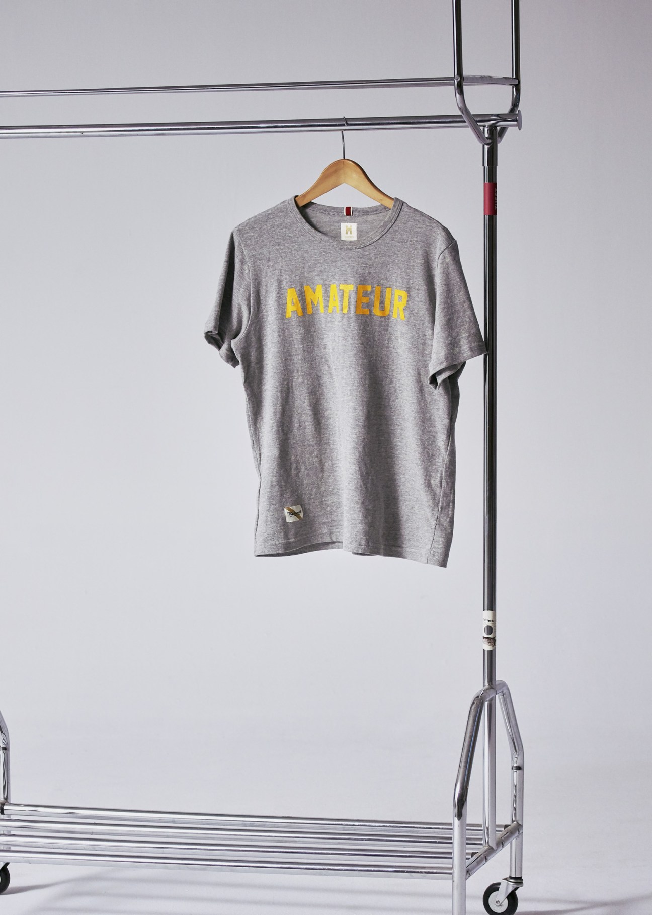 Gray T-shirt with yellow 'AMATEUR' text printed on front, hanging on wooden hanger.