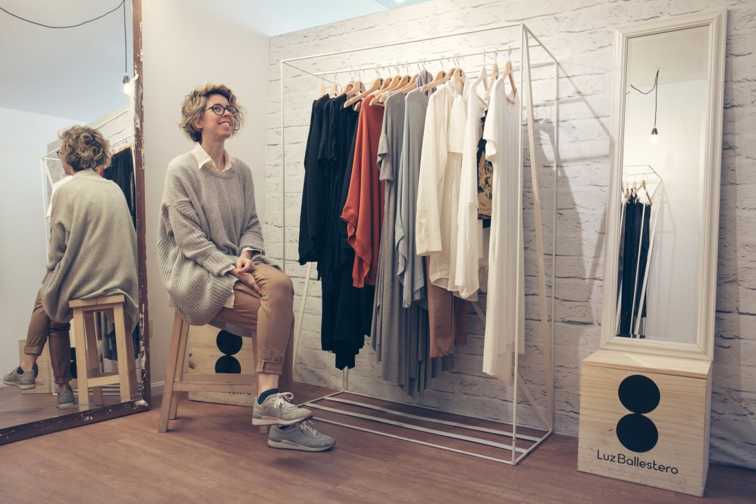 Woman in glasses sitting in a showroom displaying Luz Ballestero womenswear on metal racks.