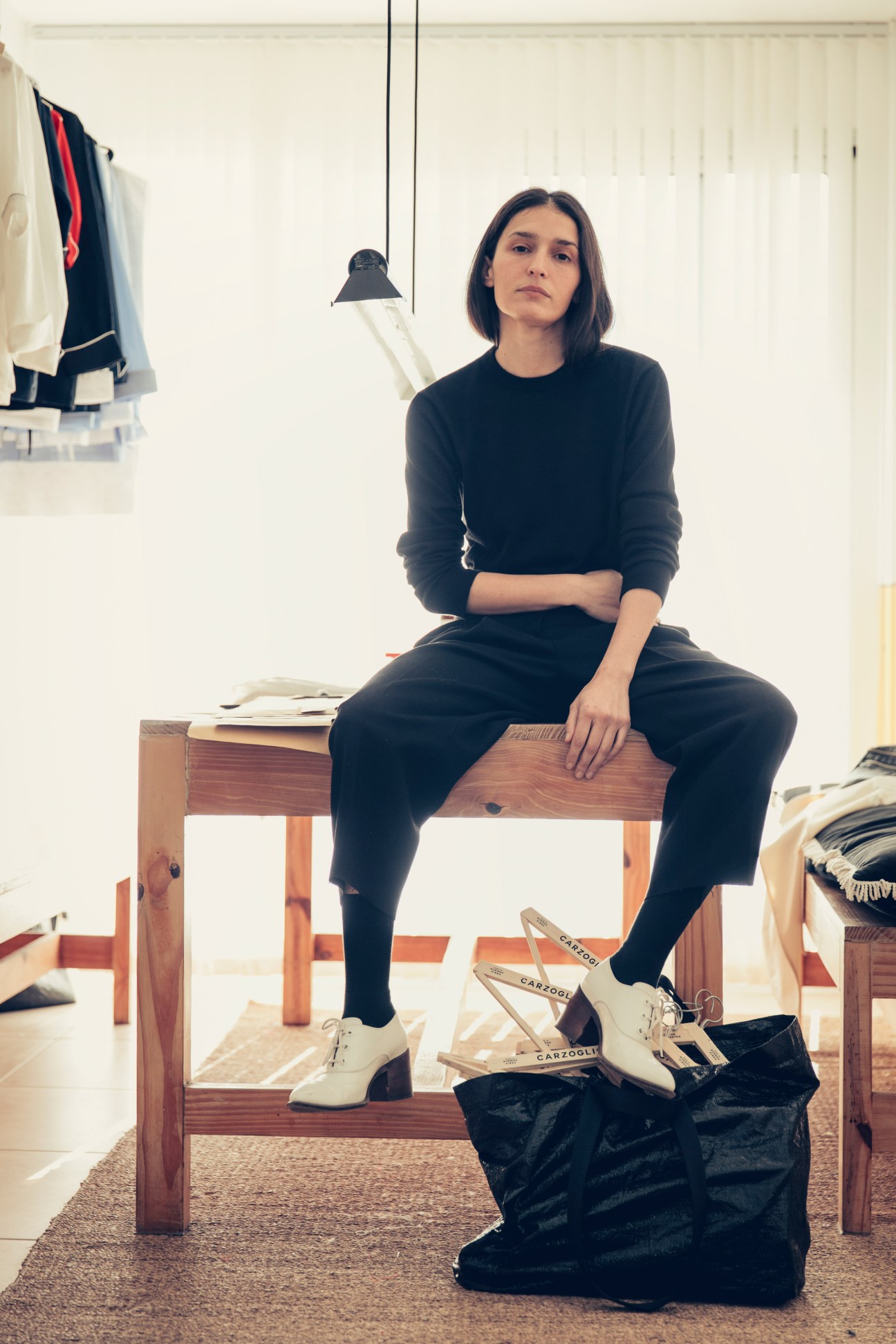 Celina Carzoglio sits on a wooden bench in her showroom wearing a black sweater and trousers, with shoes displayed below.