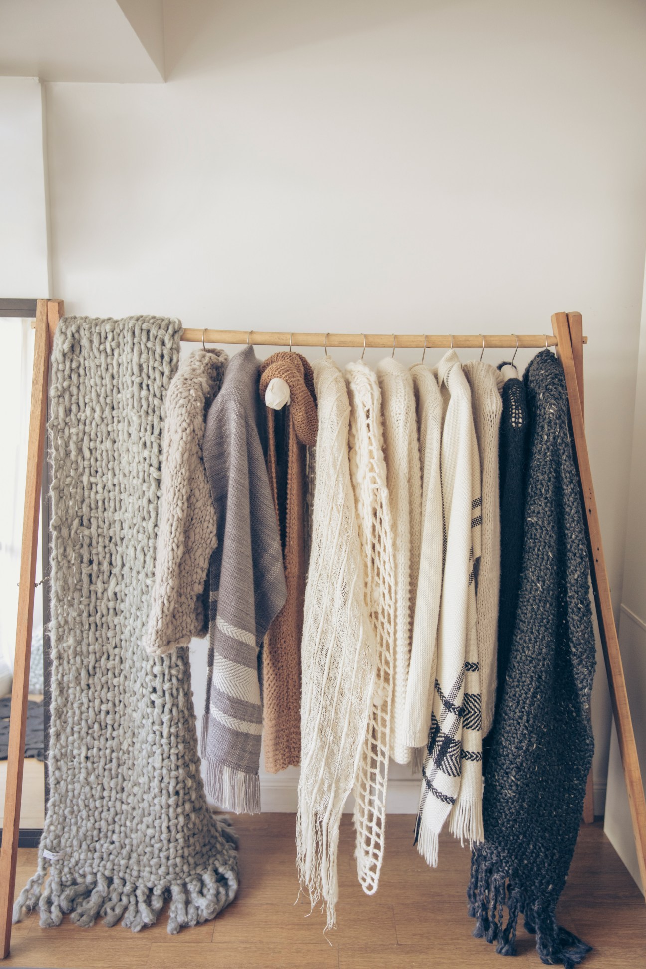 Handknitted sweaters and scarves in neutral and dark tones displayed on a wooden rack in Maydi's studio.