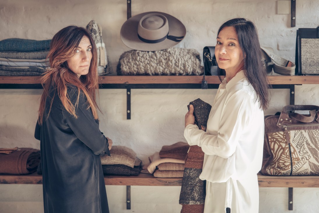 Manto Abrigos co-founders Clara de la Torre and Diana Dai Chee Chaug stand before shelving displaying handknit overcoats and textiles.
