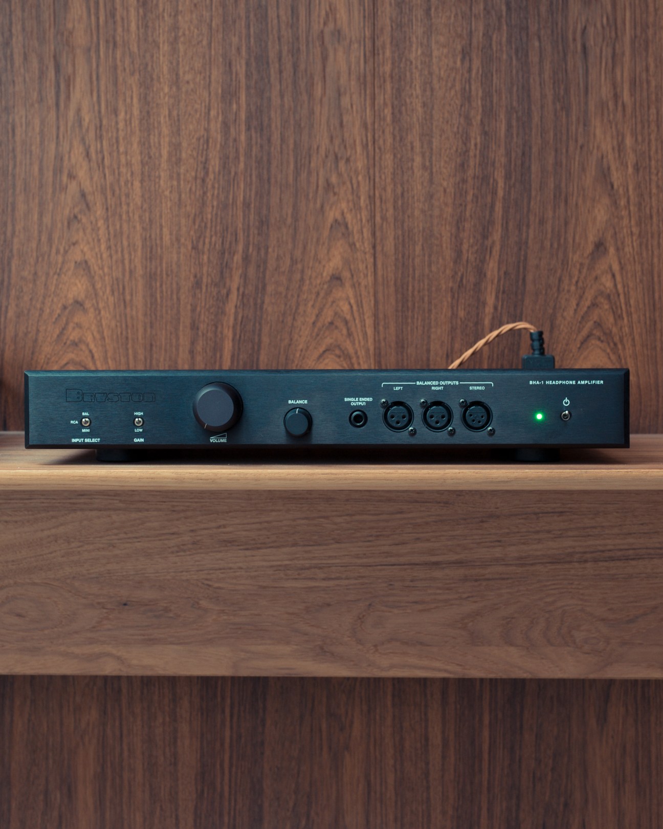 Bryston BHA1 headphone amplifier on wooden shelf against wood-paneled background.
