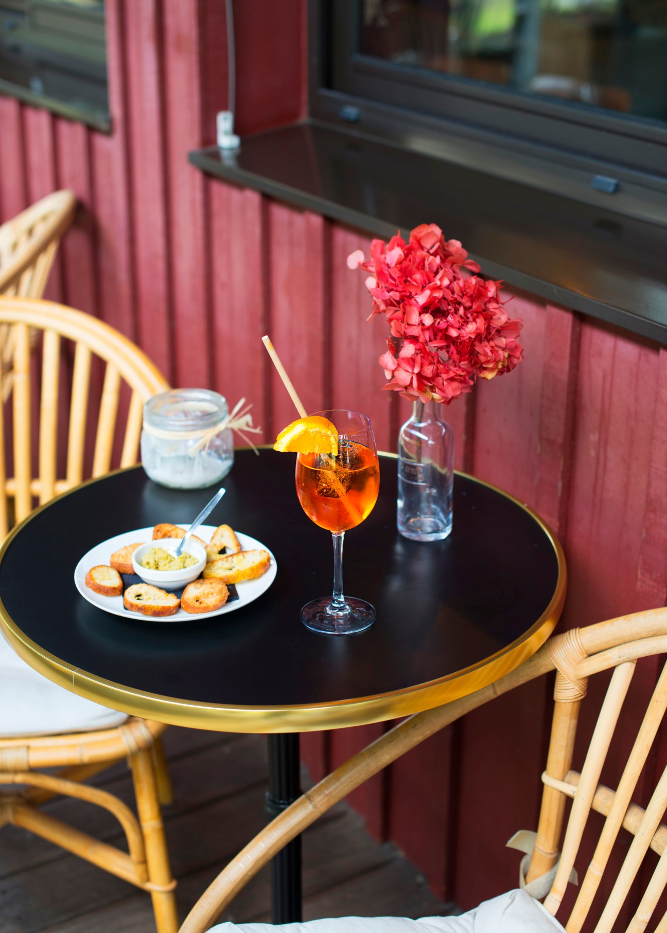 Outdoor café table with cocktail, appetizers, and red flowers against burgundy backdrop.
