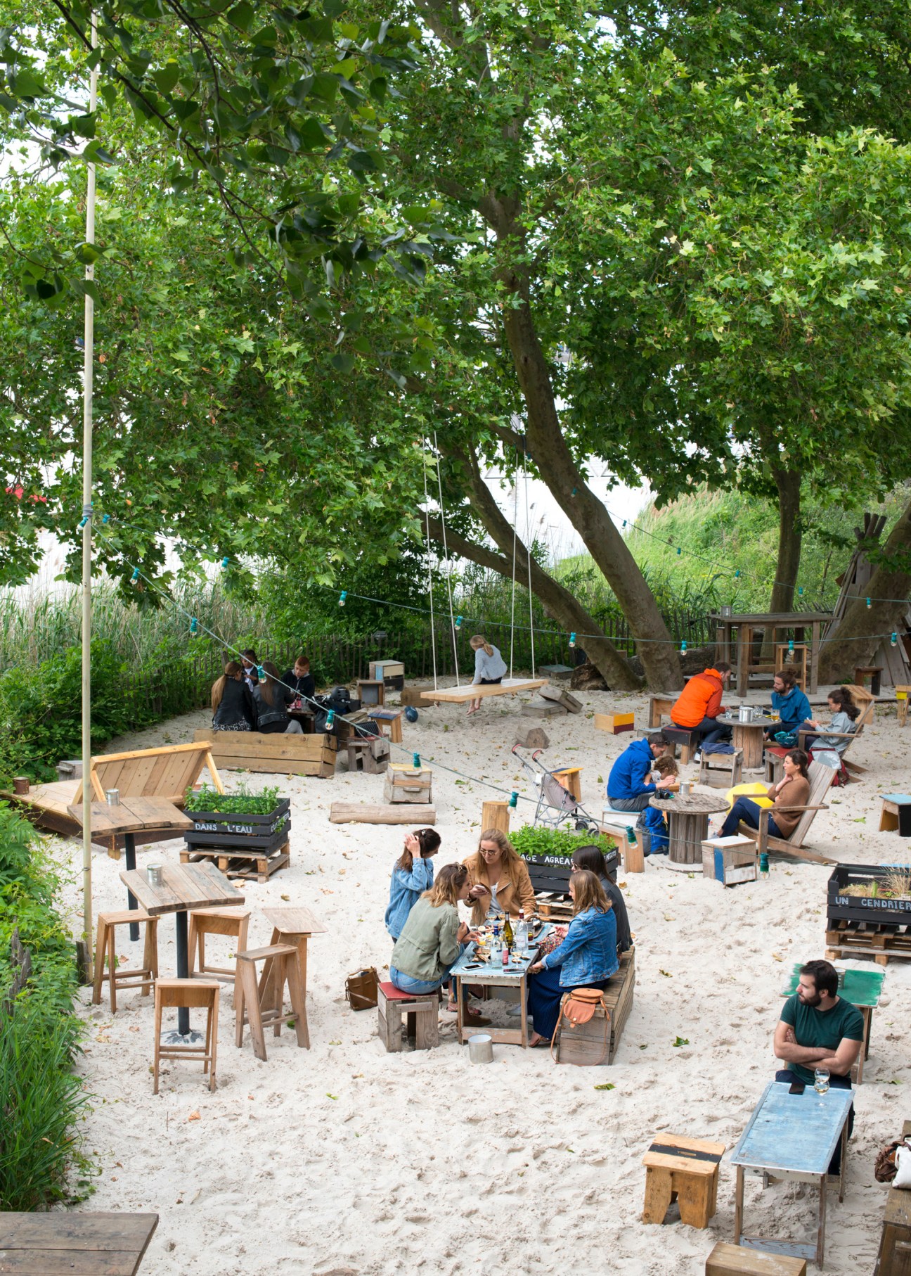 Les Chantiers de la Garonne riverside bar and brewery with sandy beach seating area, wooden furniture, and people socializing under large trees.