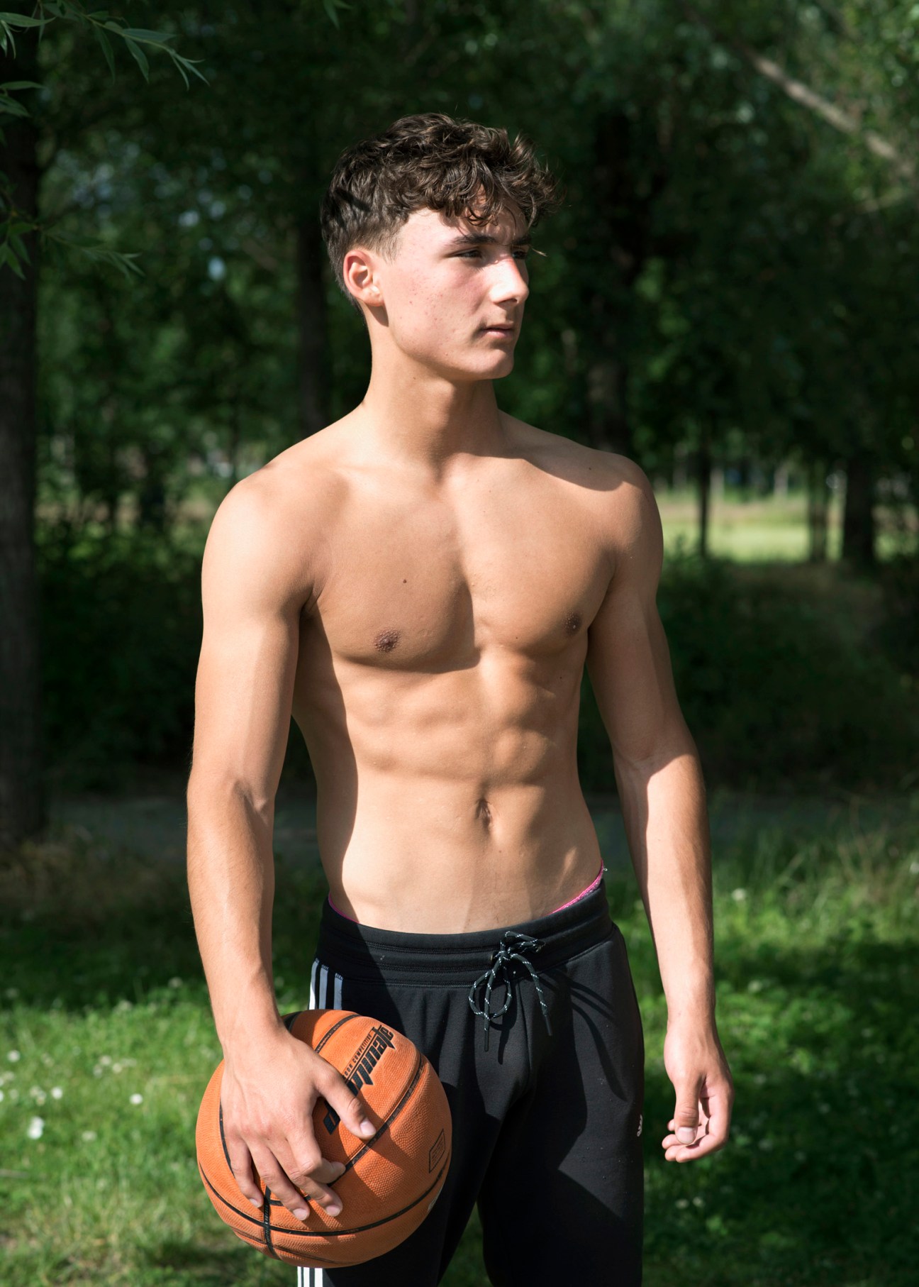 Young man holding a basketball outdoors in a green park setting.