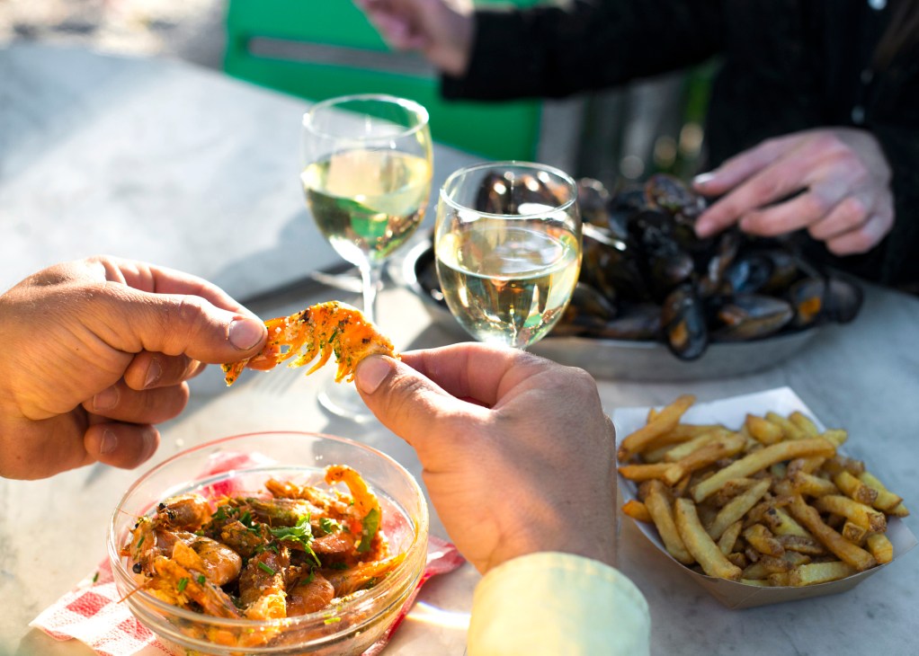 Hands holding seafood and wine glasses at an outdoor waterside meal in Bordeaux.