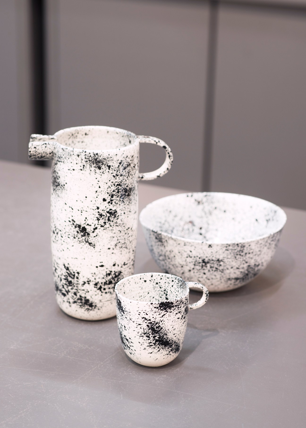 Speckled ceramic tableware set with white and black speckled jug, bowl, and cup on gray surface.