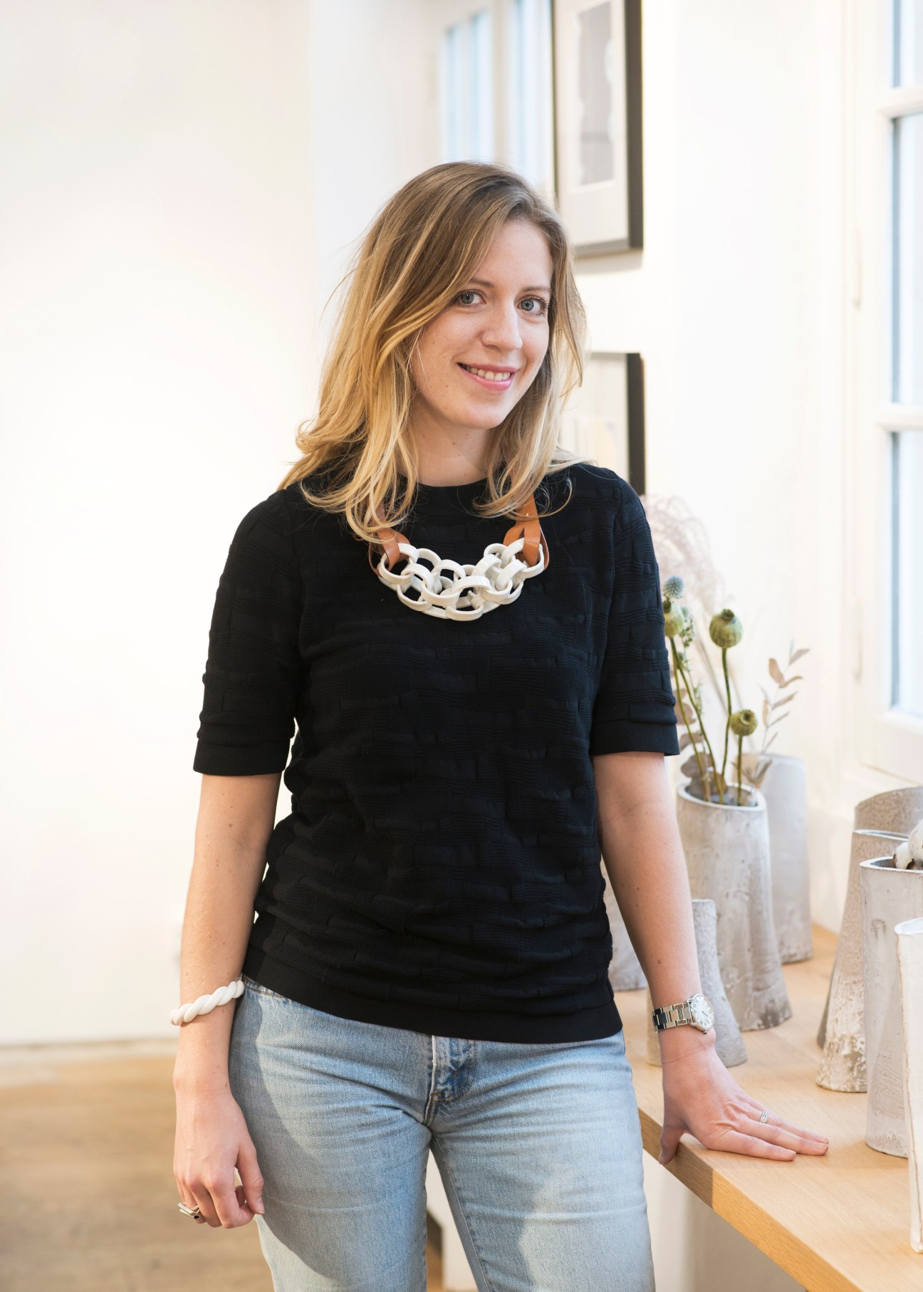 Woman with blonde hair wearing black top and blue jeans smiles in a bright interior space