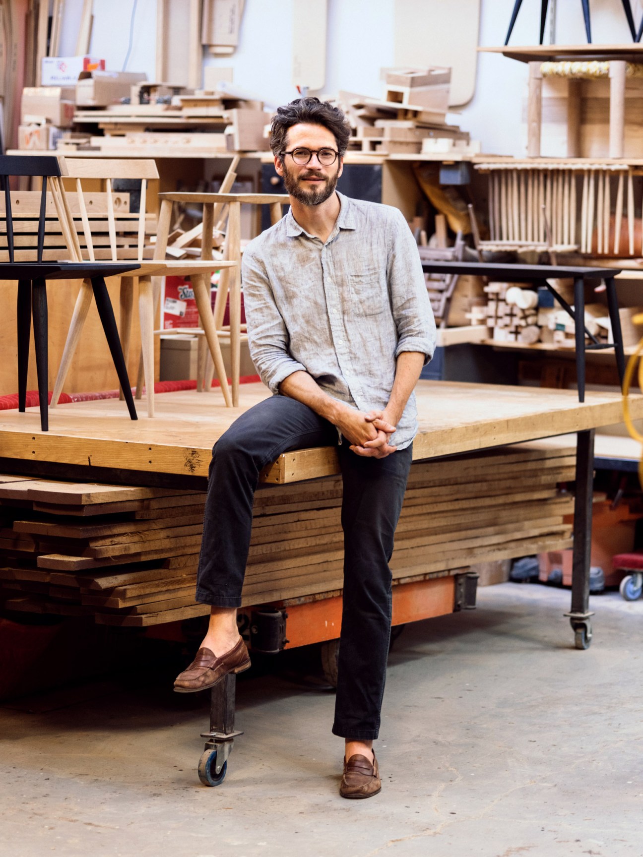 Stephen Dalrymple sitting on a wooden workbench in his furniture workshop surrounded by chairs and wooden pieces.