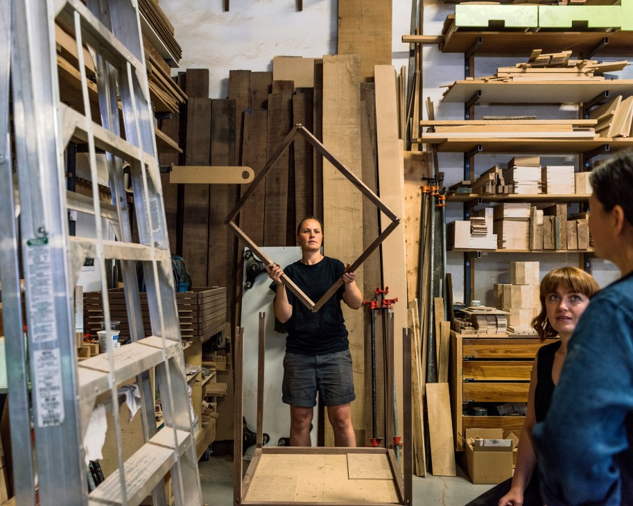Carey Jernigan holds a wooden frame in Junction Workshop studio surrounded by wood materials and shelving.
