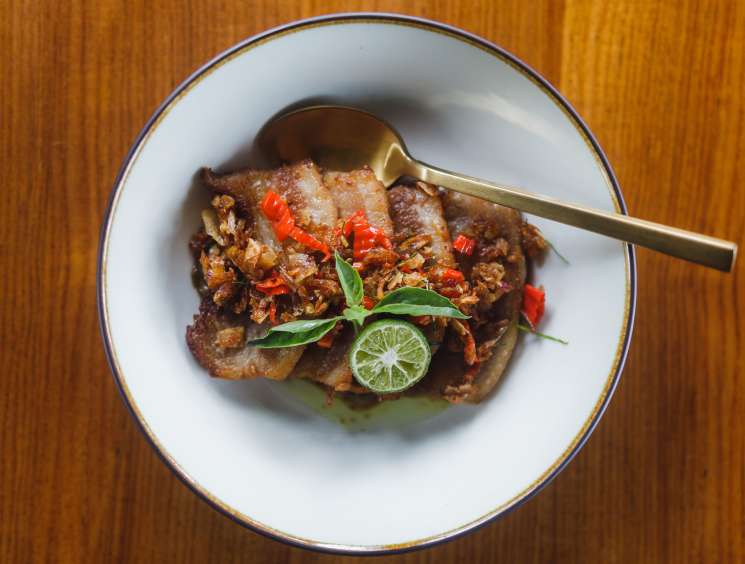 Spicy Babi Genyo pork cheek dish garnished with red chilies and lime on a white plate with brass cutlery.