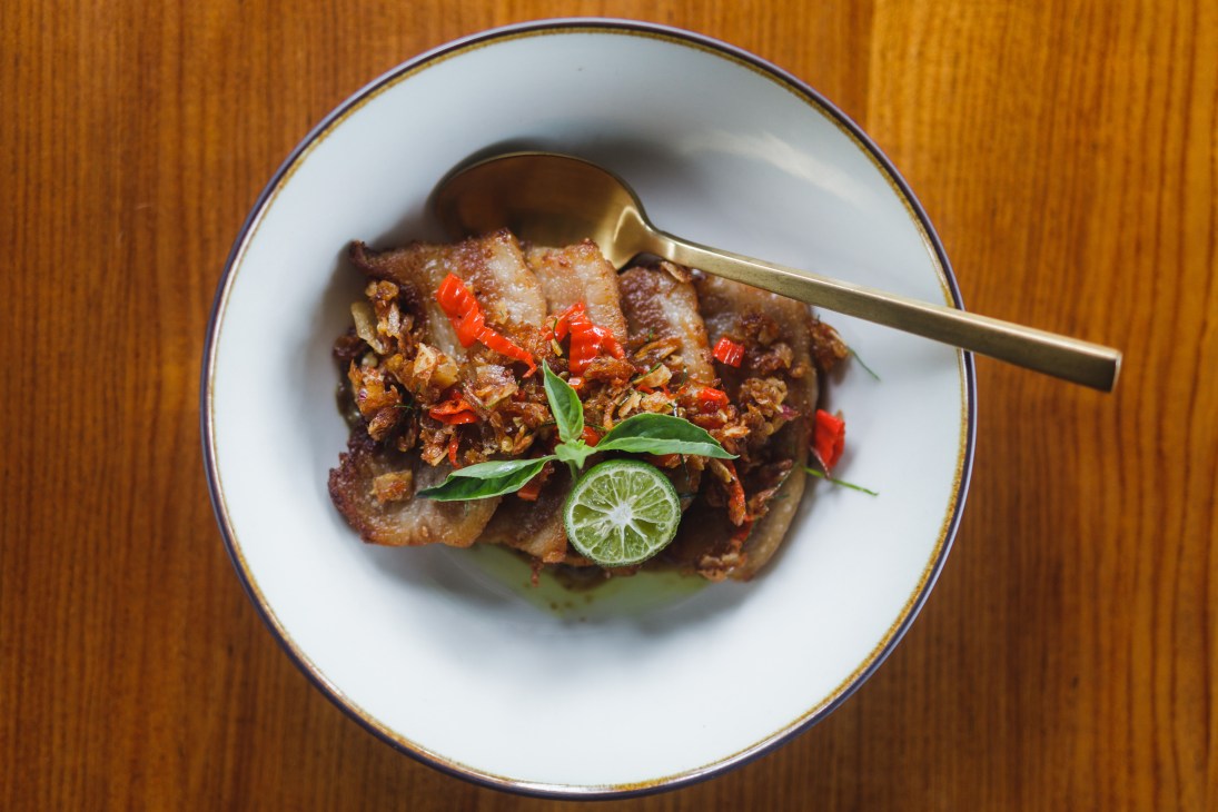 Spicy Babi Genyo pork cheek dish garnished with red chilies and lime on a white plate with brass cutlery.