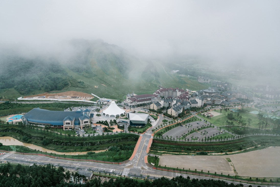 Aerial view of Alpensia Resort with Olympic venues, hotels, and surrounding forested mountains in misty conditions.