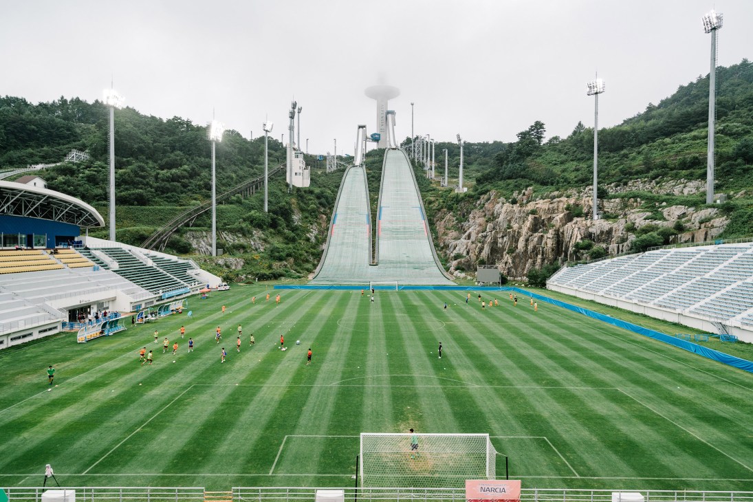 Alpensia Ski Jumping Stadium with two large ski jump ramps, sports field below, and forested mountains.