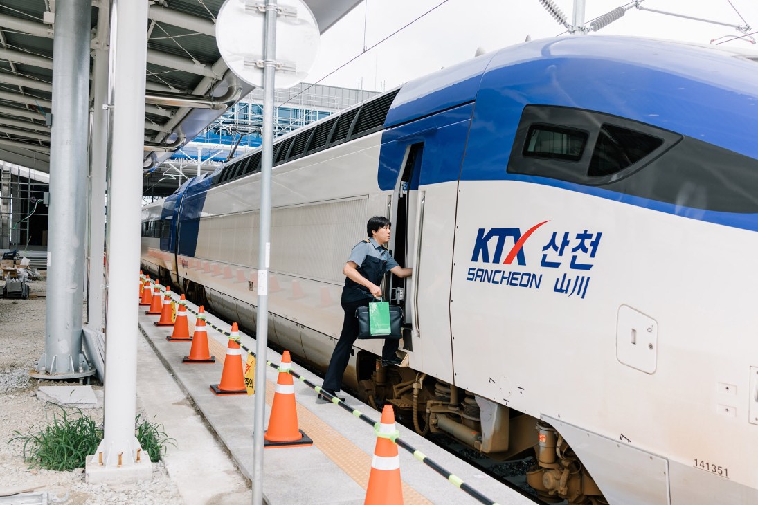 A KTX bullet train at Sancheong station with a worker boarding, surrounded by orange traffic cones during construction.