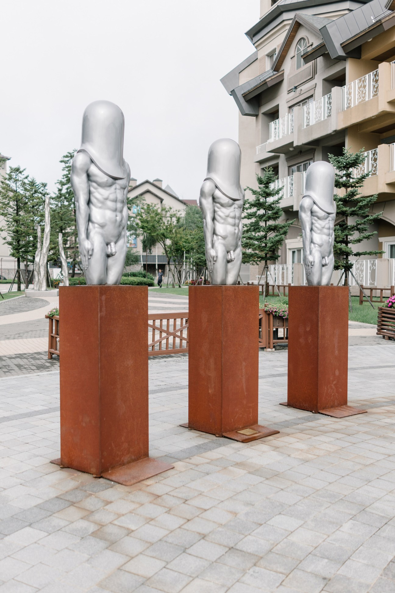 Three silver human statues on red-brown pedestals in a plaza at Alpensia Resort, with resort buildings and landscaping in background.