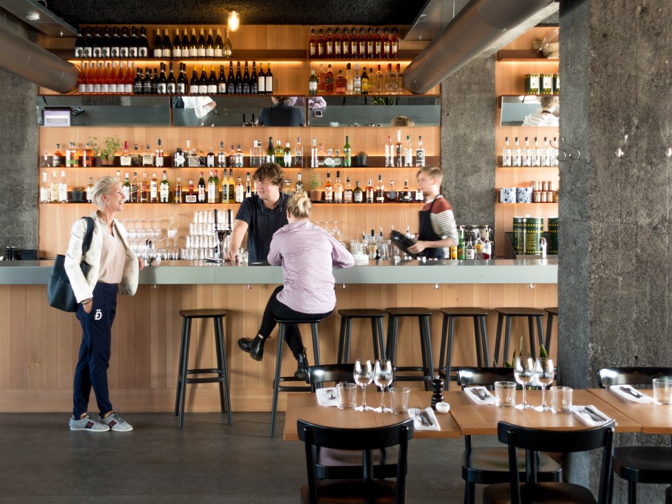 Modern restaurant interior with concrete columns, wooden bar, extensive bottle display, and customers at bar stools