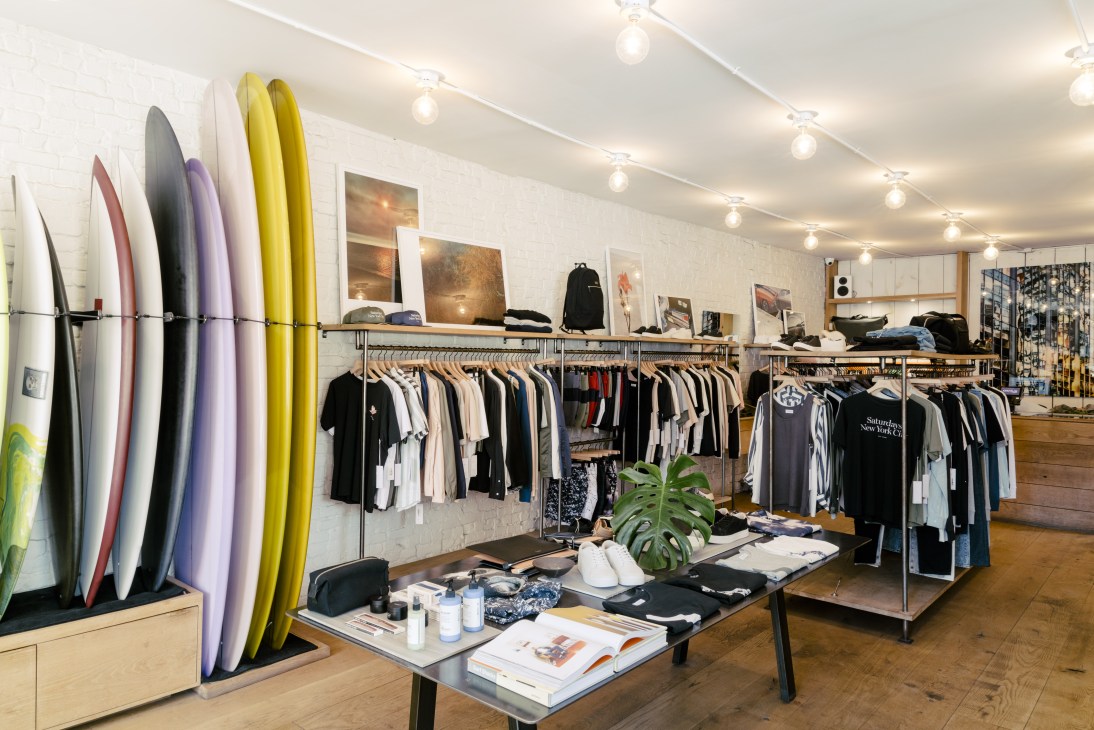 Interior of Saturdays NYC store on Crosby Street with colorful surfboards on left wall, clothing racks, and white brick walls.