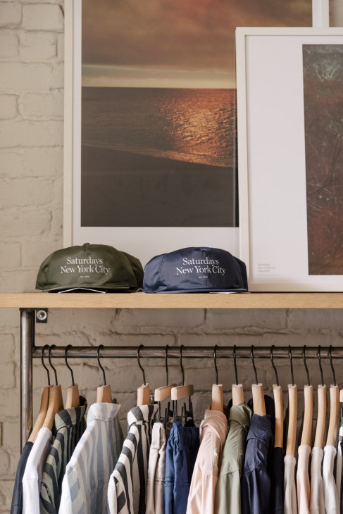 Two Saturdays NYC baseball caps in olive and navy displayed on a wooden shelf above hanging clothing.