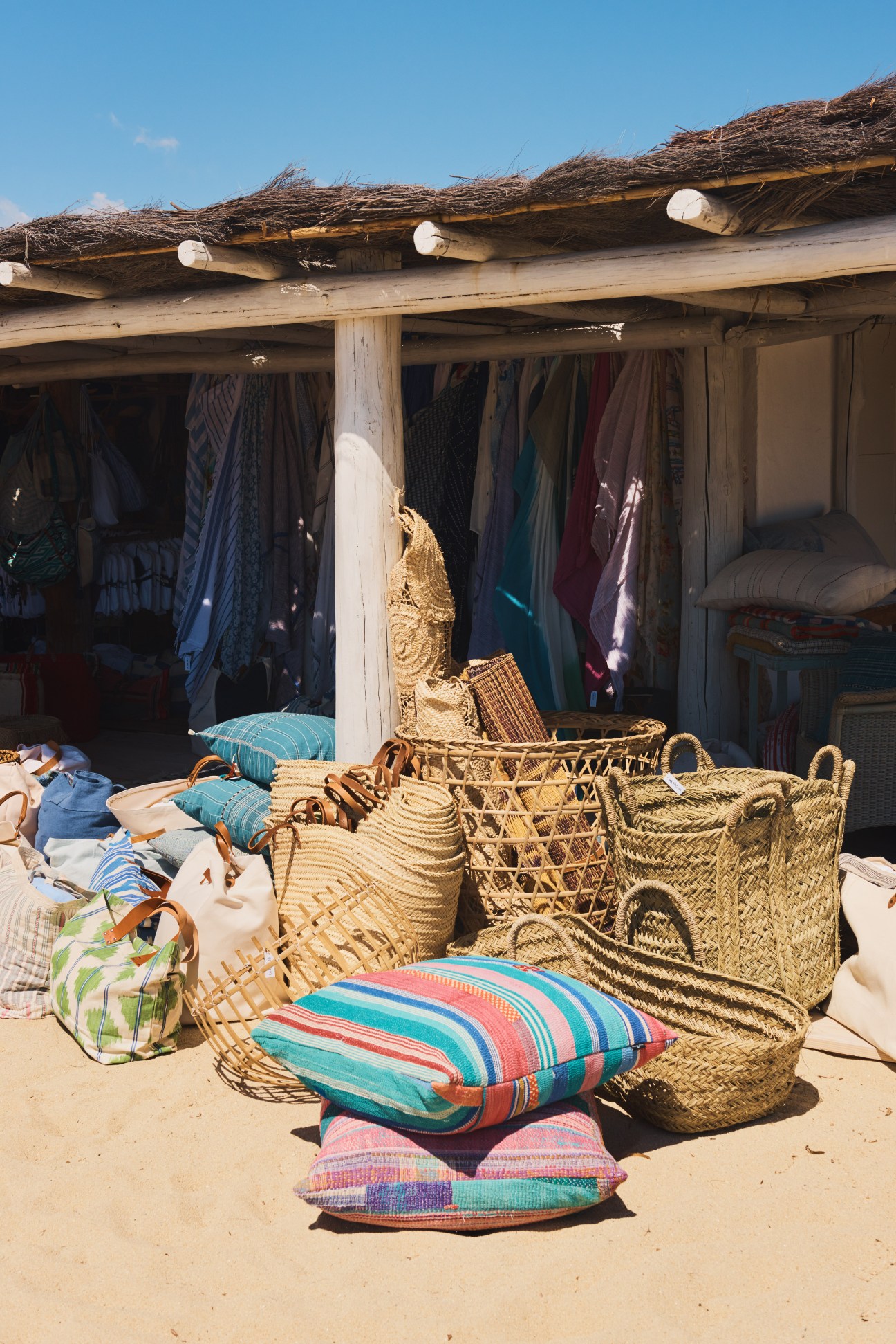 Club 55 boutique merchandise displayed on sand including woven baskets, striped pillows, and colorful clothing hanging from a thatched roof structure.