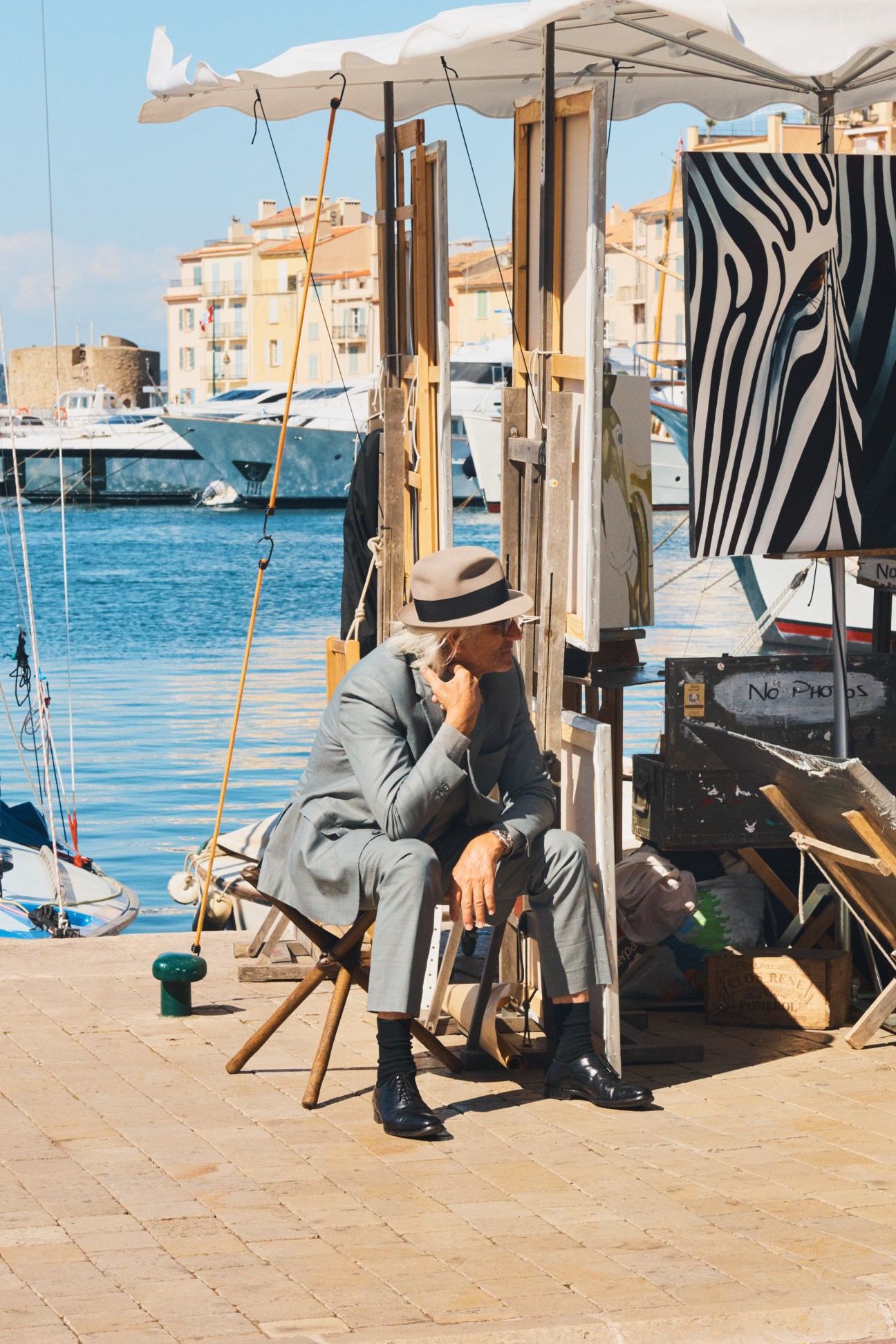 Man in grey suit and straw hat sitting on waterfront with boats and Mediterranean buildings in background.