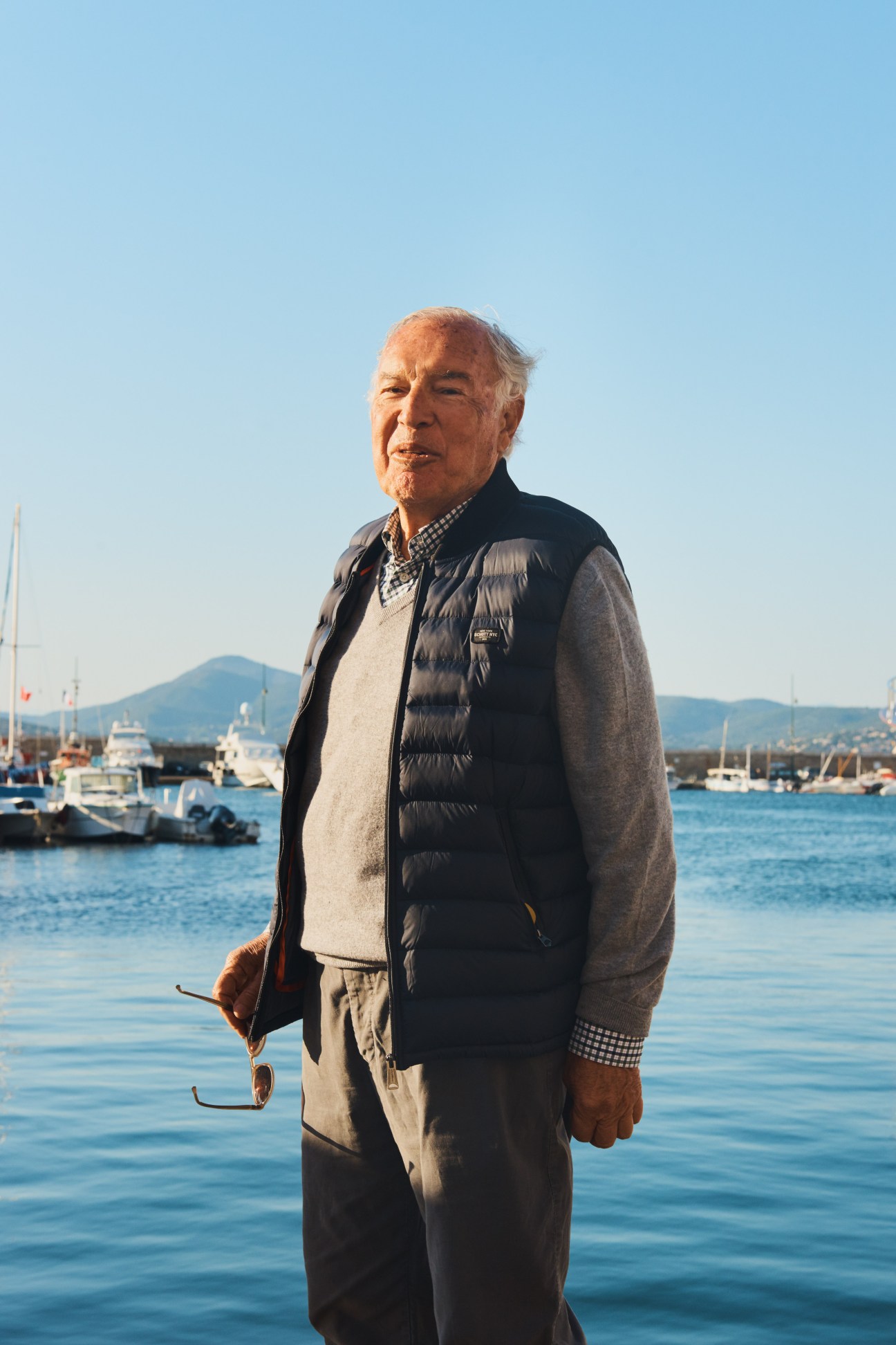 Elderly man in navy puffer vest and checkered shirt standing by St Tropez harbour with boats behind him.