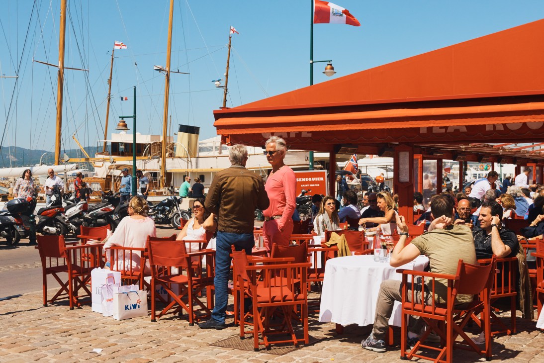 Senequier waterfront café with red chairs and umbrellas, sailboats and harbor in background.