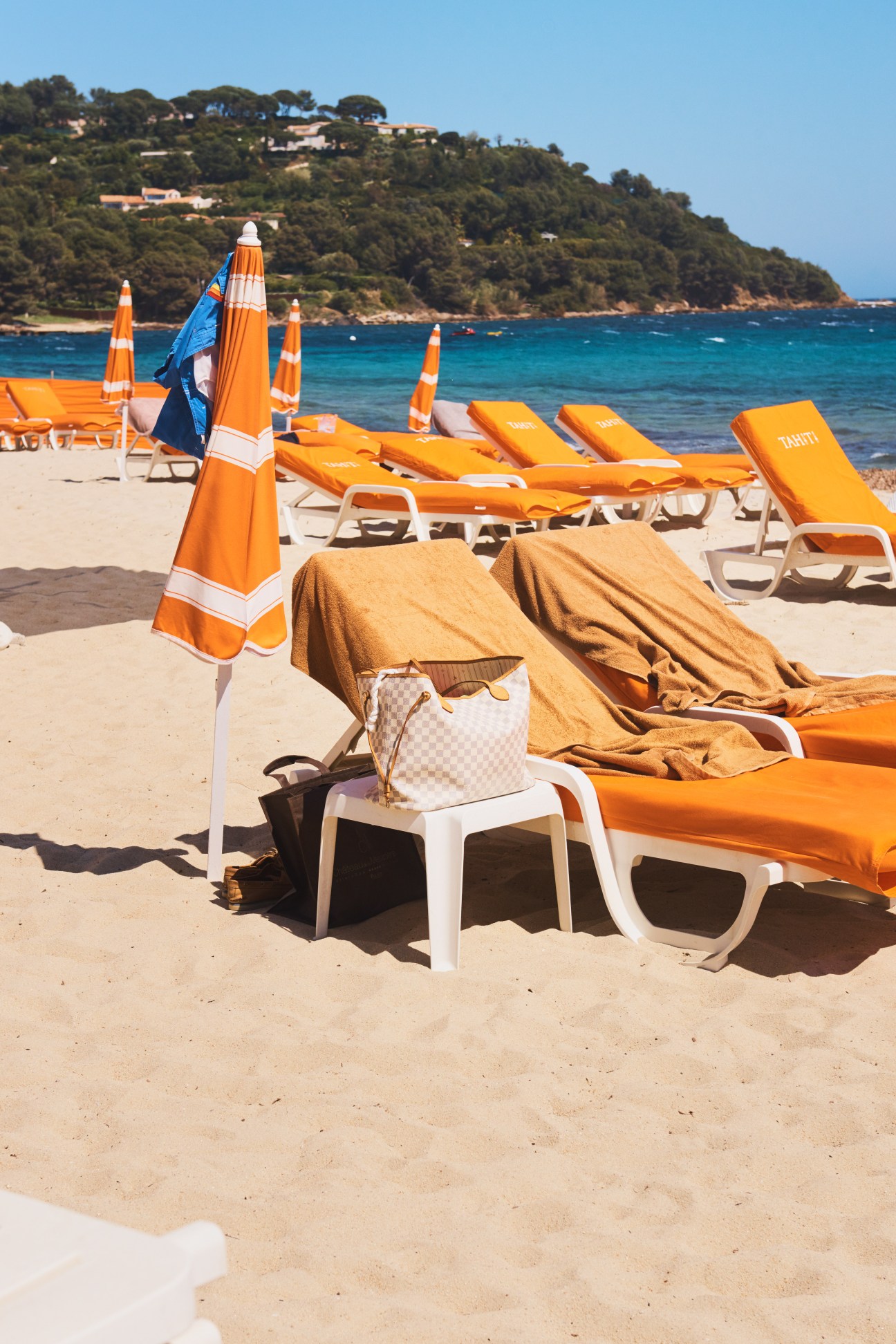 Tahiti beach club with orange umbrellas, loungers and towels on sandy beach overlooking turquoise Mediterranean water.