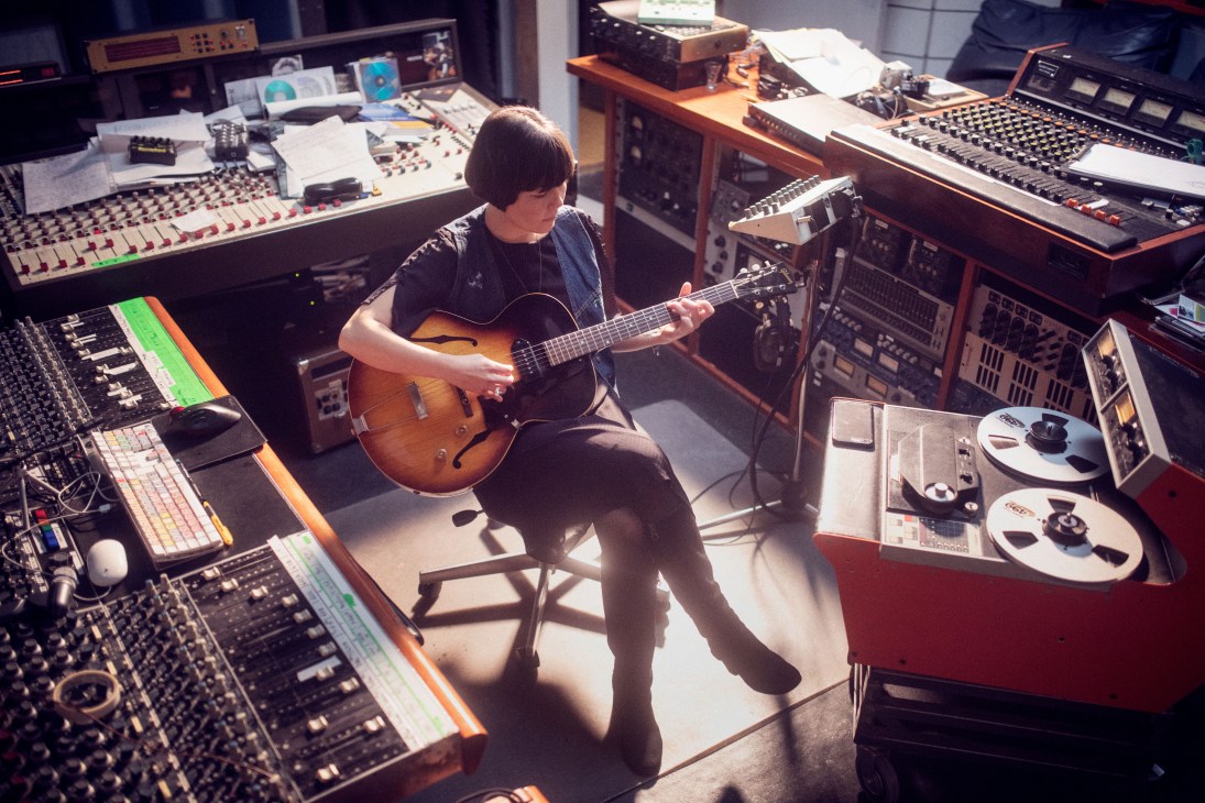 Sumie sits playing acoustic guitar in a Gothenburg recording studio surrounded by mixing equipment and synthesizers.