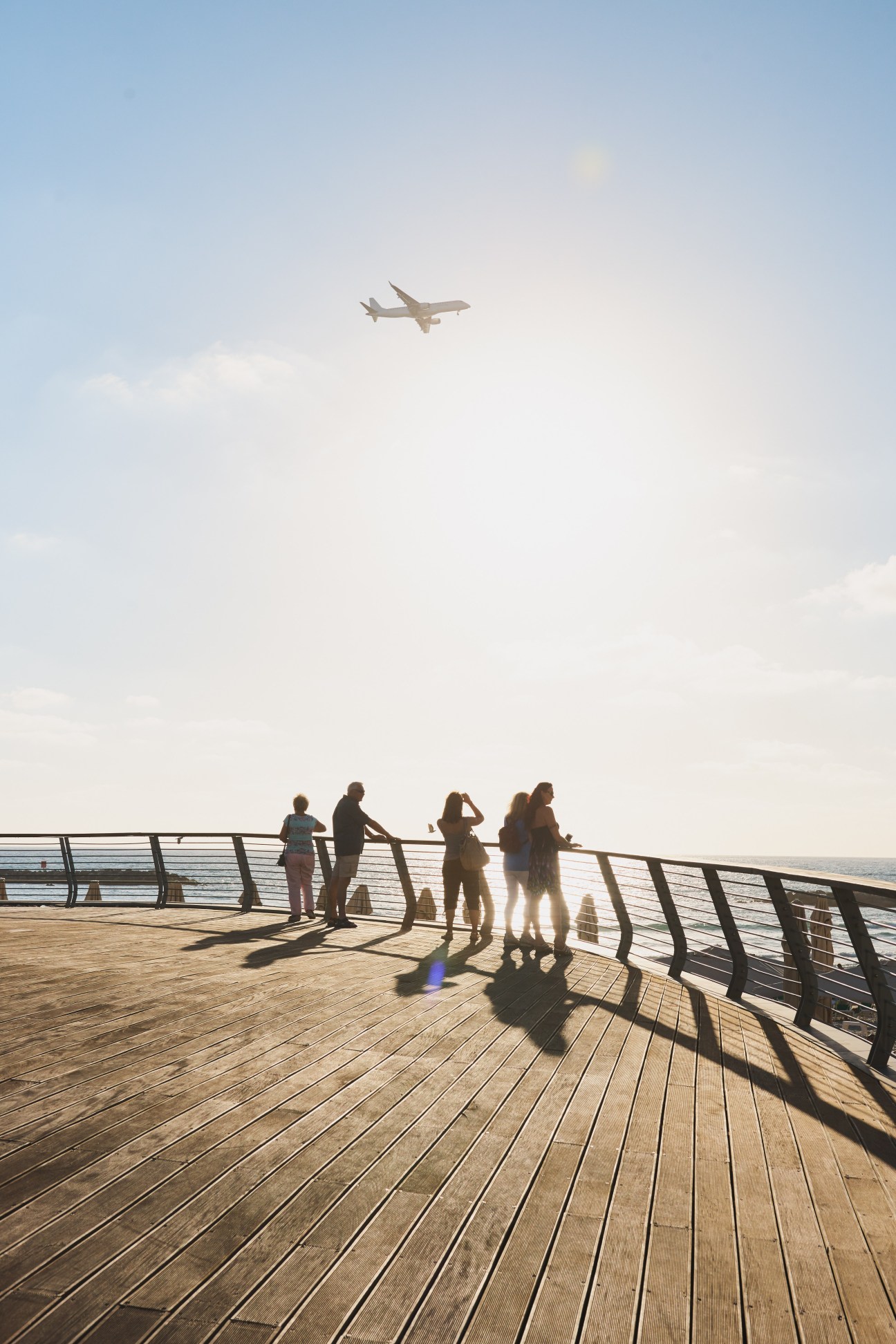 People standing on a wooden boardwalk by the sea with an airplane flying overhead.