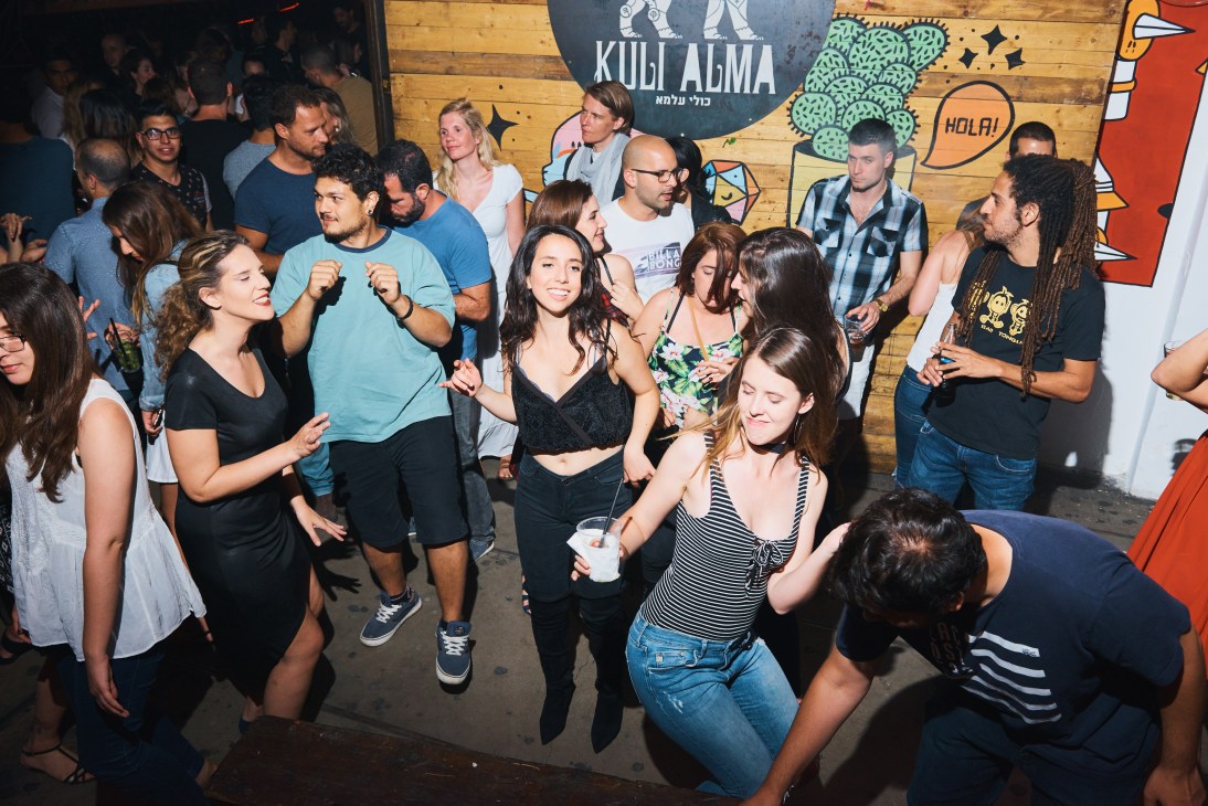 Diverse crowd dancing together at Kuli Alma nightclub in Tel Aviv with colorful street art murals on wooden walls.