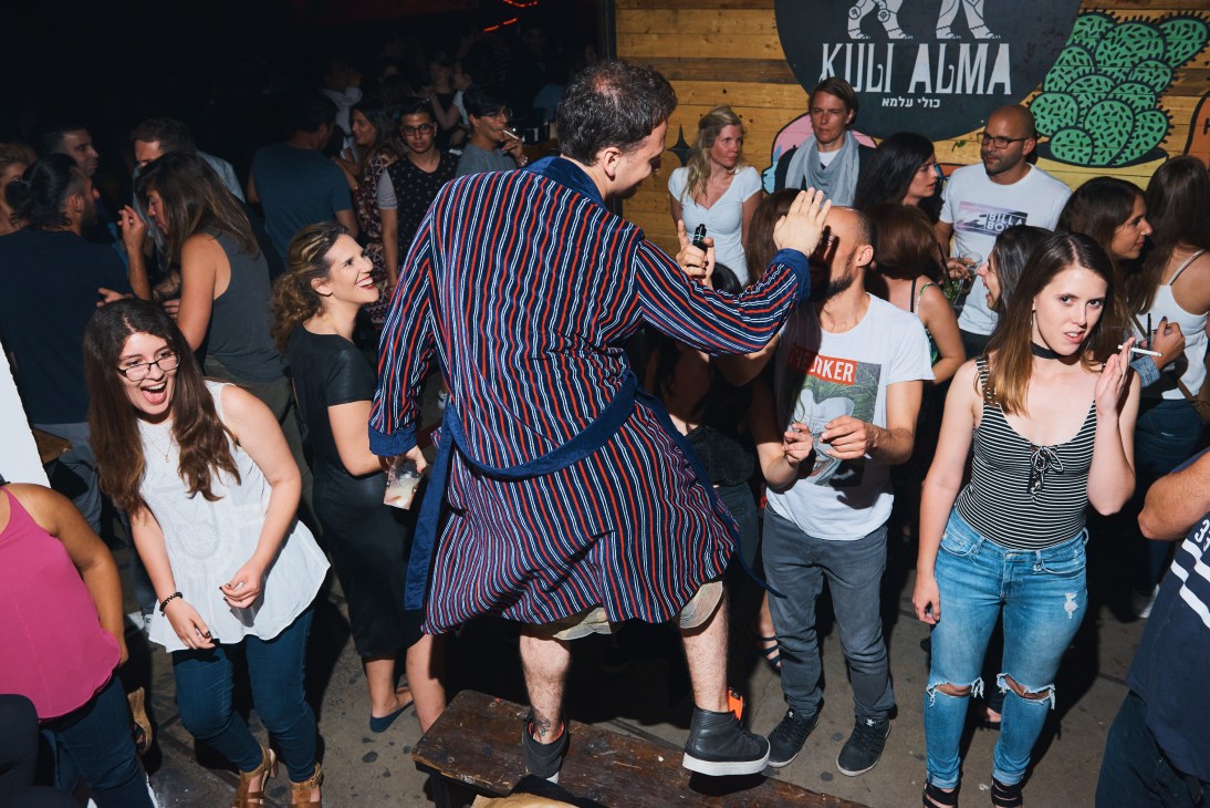 Man in red and blue striped shirt dancing at Kuli Alma nightclub surrounded by smiling guests.