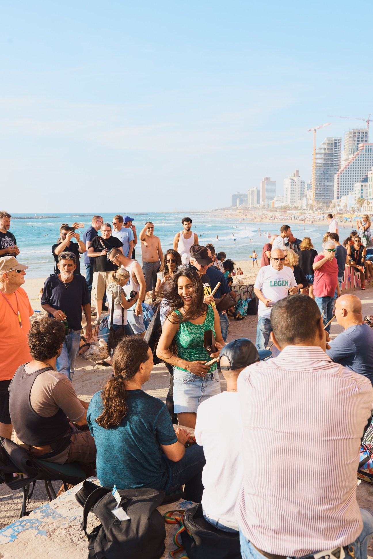 People dancing and gathering on a Tel Aviv beach with high-rise buildings and Mediterranean Sea in the background.