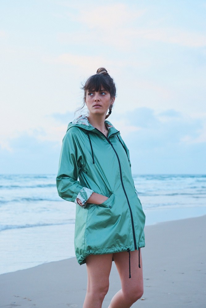 Woman in turquoise jacket standing on beach at dusk looking toward the horizon.