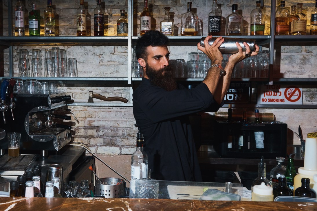 Bearded bartender in black shirt preparing a cocktail behind a bar with shelves of spirits and glasses.