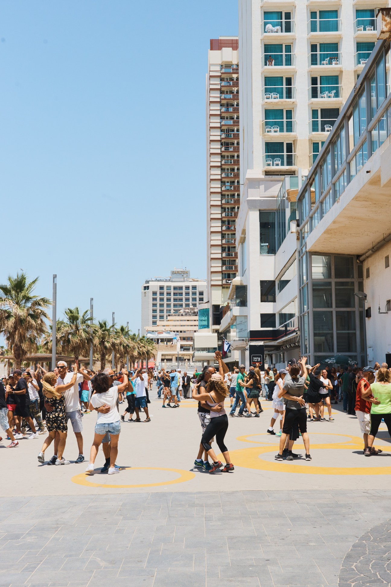 Crowds of people dancing in a Tel Aviv plaza surrounded by modern residential buildings on a sunny day.