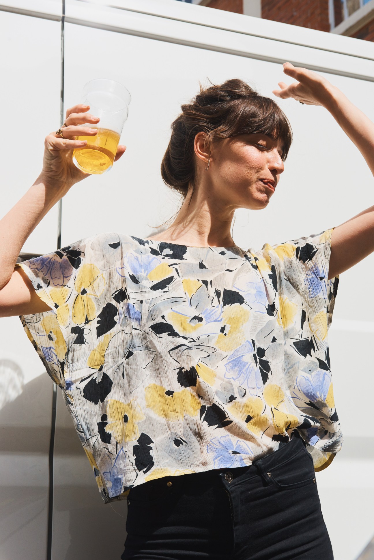Woman holding a glass of beer with arms raised in a celebratory pose against a white wall.