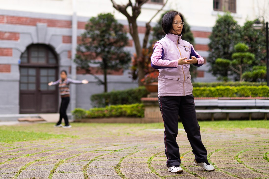Woman in pink jacket practicing tai chi in a courtyard garden of a brick building.