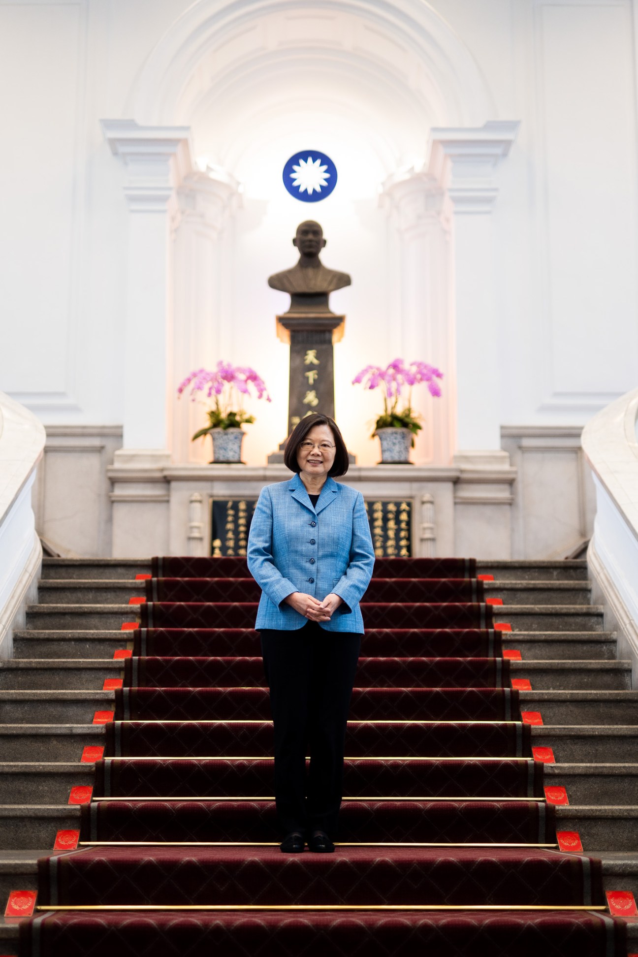 Taiwan's President Tsai Ing-wen stands on grand red-carpeted stairs before a bronze bust of Dr Sun Yat-sen, wearing a blue jacket.