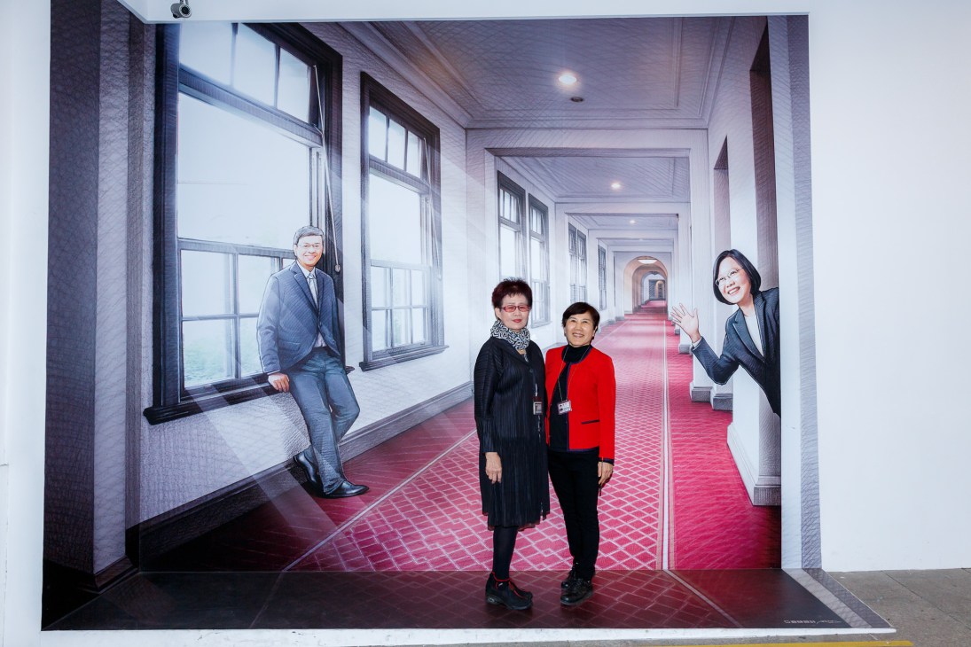 Two women pose in front of a life-size cardboard cutout of President Tsai Ing-wen and her vice-president in a corridor.