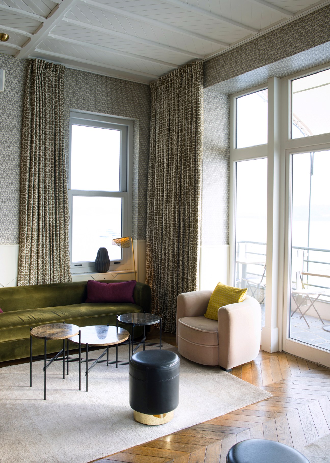Modern hotel lounge with olive green sofa, cream armchair, and floor-to-ceiling windows overlooking a balcony.