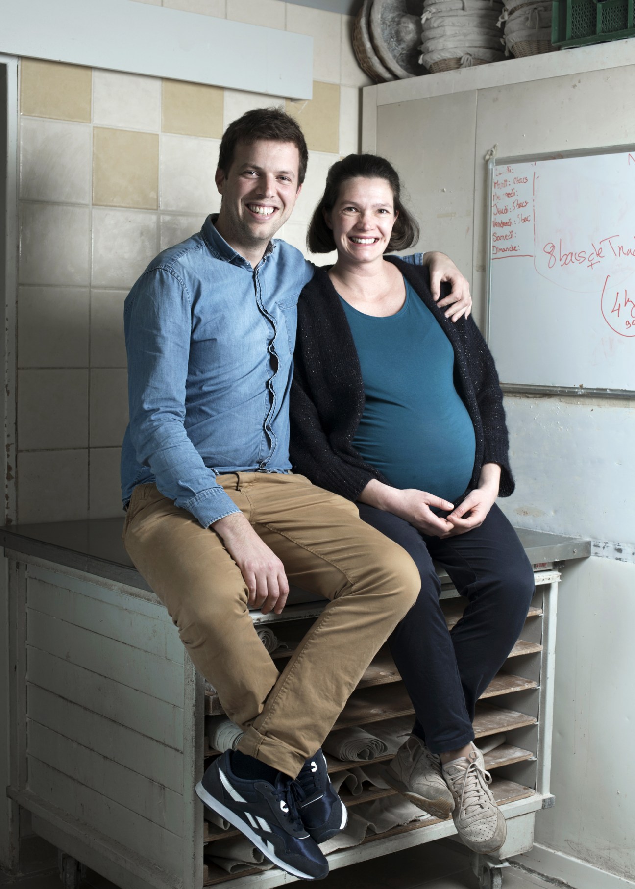 Matthieu and Caroline Gailly sit together on a wooden bench in their biscuit factory kitchen, smiling at the camera.