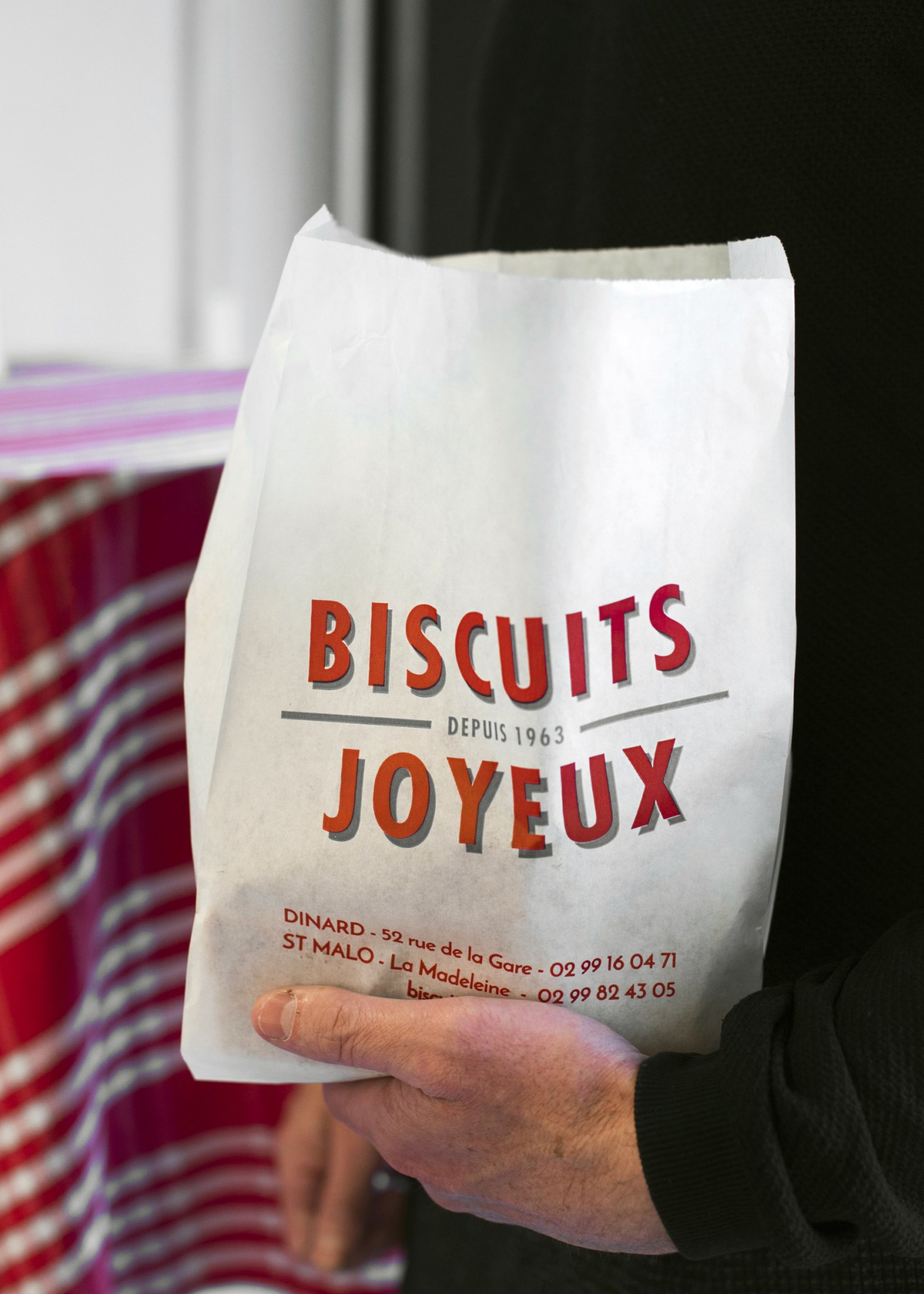 Hand holding a white paper bag printed with red text reading 'Biscuits Joyeux' and 'Depuis 1963'.
